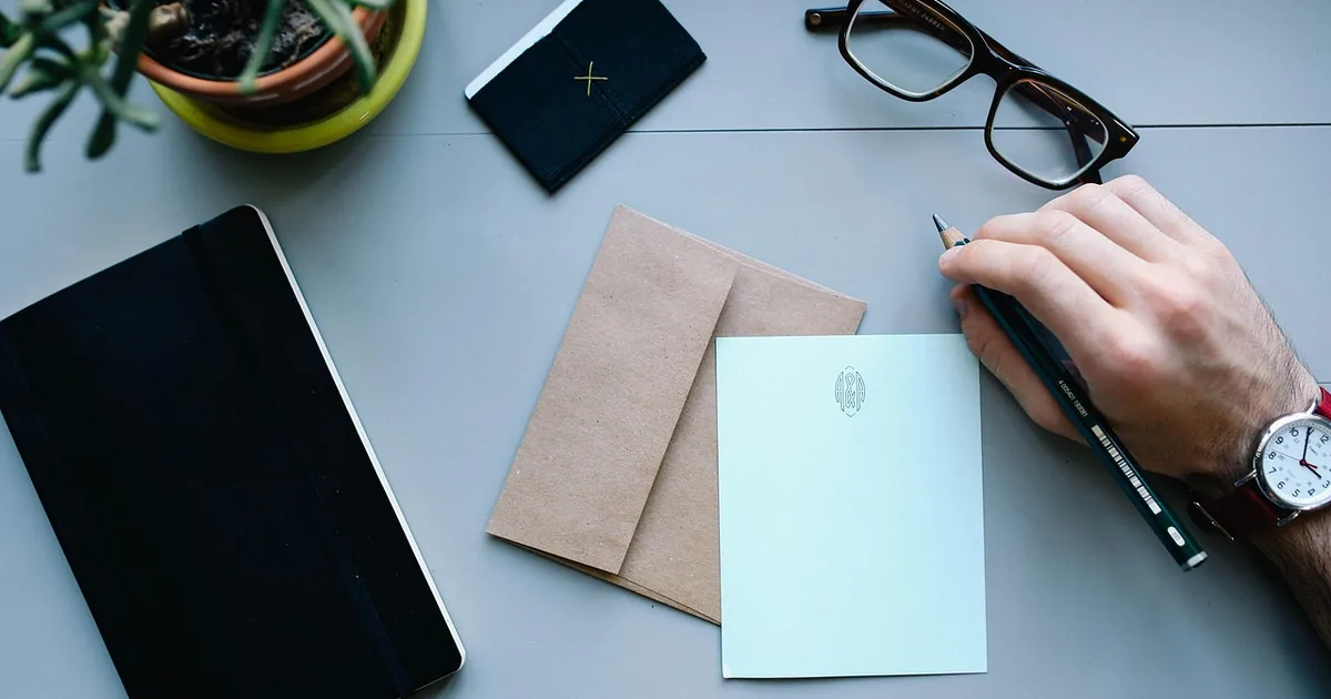 Bureau vu de dessus : une main rédige un courrier, à côté d’une enveloppe kraft et d’un carnet.