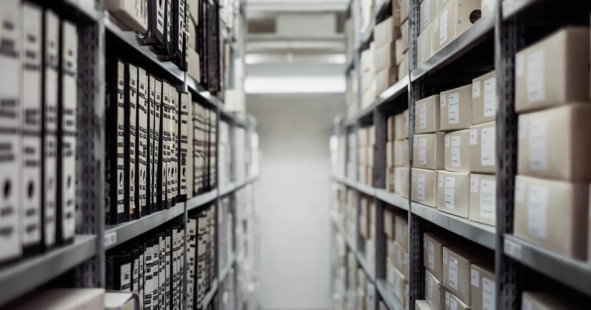 Couloir d’archives bordé de deux rangées d’étagères se faisant face, chargées de classeurs et de boîtes.